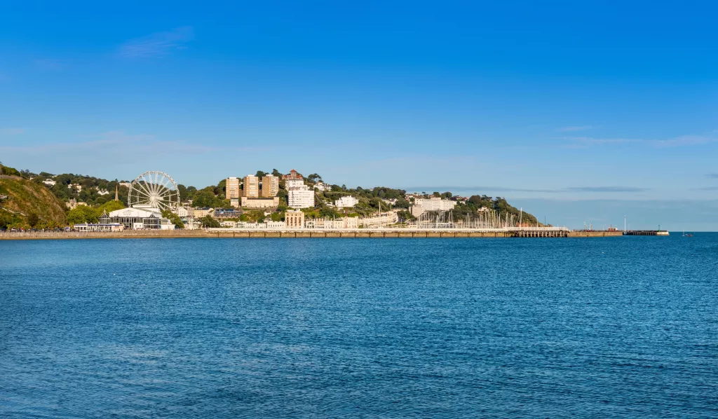 Scenic view of Torquay seafront with calm blue water, the town’s big wheel, marina, and hillside buildings visible under a clear sky.