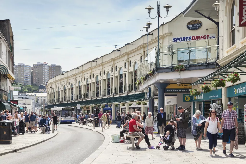 Busy shopping street in Torquay with people walking, sitting, and shopping outside Fleet Walk, with visible shops including Poundland and Sports Direct